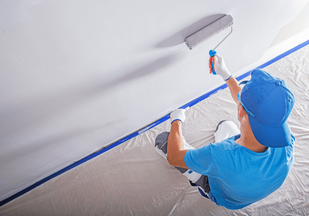 A person painting a wall with a roller while wearing gloves and a blue cap, with a drop cloth on the floor.